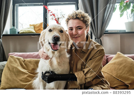 Portrait of young woman with prosthetic arm embracing her lovely golden retriever and looking at camera while sitting on sofa in living room Portrait of young woman with prosthetic arm embracing her lovely golden retriever and looking at camera while sitting on sofa in living room 88393295