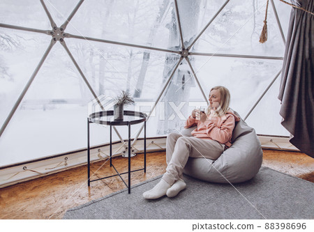 A woman is sitting in a geo dome glamping tent in a bag chair and looking out at the winter snow-covered nature and drinks coffee. 88398696