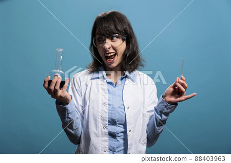 Creepy looking biochemistry specialist holding glass vials filled with chemical liquid substance samples on blue background. Microbiology crazy and lunatic looking expert having chemistry jars. Creepy looking biochemistry specialist holding glass vials filled with chemical liquid substance samples on blue background. Microbiology crazy and lunatic looking expert having chemistry jars. 88403963