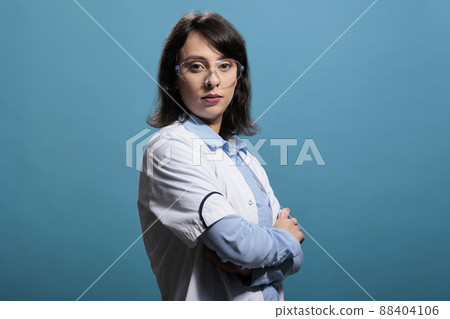 Biochemist scientist expert wearing lab coat and protective goggles, standing on blue background with crossed arms. Chemistry specialist wearing protective glasses and white coat while standing on 88404106