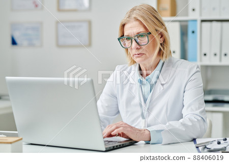 Serious mature blond female doctor typing on laptop keyboard in office 88406019