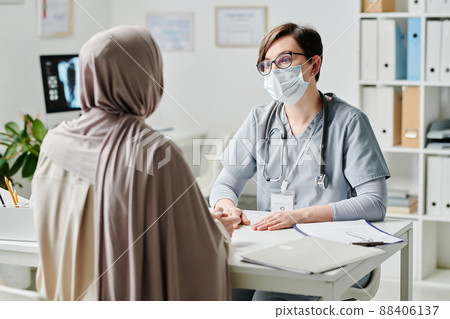 Contemporary doctor in protective mask talking to female patient in hijab 88406137