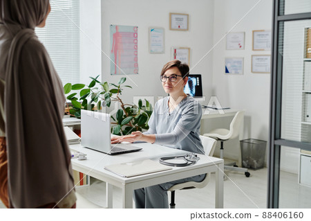 Young smiling female clinician inviting Muslim patient sit in front of her Young smiling female clinician inviting Muslim patient sit in front of her 88406160