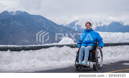 A happy woman dressed in a blue coat sits in a wheelchair on a point view and looking at the snow-capped mountains.  88408585