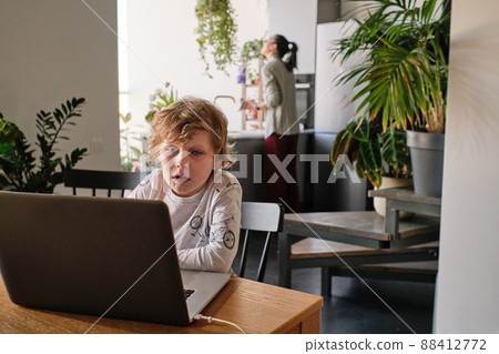 Little boy sitting at table in kitchen and using laptop, he studying online or playing game Little boy sitting at table in kitchen and using laptop, he studying online or playing game 88412772