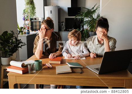 Upset homosexual mothers trying to teach their naughty son sitting at table with books in kitchen 88412805