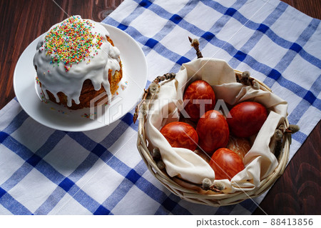 Easter cake in white glaze with multicolored caramel topping and Easter eggs in the shape of a cross with a pattern of terracotta lace lie in a basket on a table. Easter holiday traditional foods Easter cake in white glaze with multicolored caramel topping and Easter eggs in the shape of a cross with a pattern of terracotta lace lie in a basket on a table. Easter holiday traditional foods 88413856