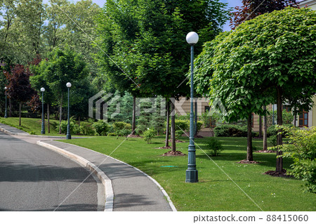 an asphalt road with a stone curb and a tarmac pedestrian sidewalk near a park with trees and bushes sprinkled with mulch on a green lawn on a sunny summer day. 88415060