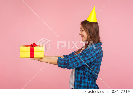 Take it, this gift for you. Side view of happy brunette girl with party cone on head giving present box and smiling, congratulating on birthday holiday. indoor studio shot isolated on pink background 88417219