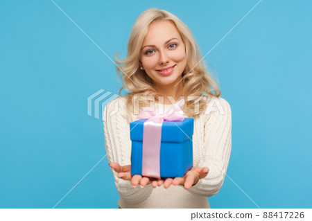Cute positive blond woman in knitted sweater holding in hands blue giftbox with ribbon bow, making present, surprise. Indoor studio shot isolated on blue background 88417226