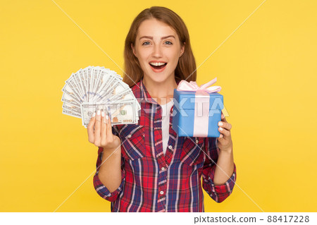 Surprising present, money bonuses. Portrait of amazed excited ginger girl in casual checkered shirt holding gift box and dollars, shocked by cashback. indoor studio shot isolated on yellow background Surprising present, money bonuses. Portrait of amazed excited ginger girl in casual checkered shirt holding gift box and dollars, shocked by cashback. indoor studio shot isolated on yellow background 88417228