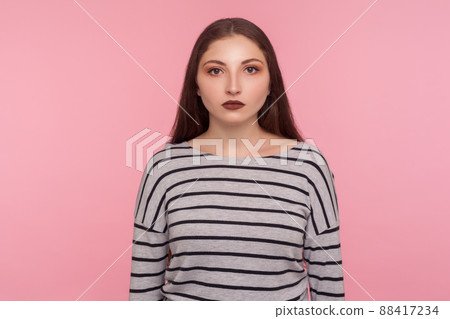 Portrait of serious calm young woman in striped sweatshirt looking at camera with unsmiling earnest expression, feeling confident focused self-assured. indoor studio shot isolated on pink background 88417234