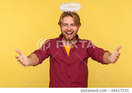 Come into my arms. Funny amiable bearded guy with saint nimbus over head stretching hands to camera and smiling friendly, going to embrace, share love. indoor studio shot isolated on yellow background 88417235