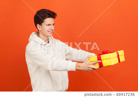 Profile portrait of smiling young man in casual sweatshirt with hood holding out present box, express delivery service. Indoor studio shot isolated on orange background Profile portrait of smiling young man in casual sweatshirt with hood holding out present box, express delivery service. Indoor studio shot isolated on orange background 88417236