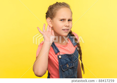 What. I can't hear. Portrait of curious attentive little girl in denim overalls holding hand near ear and listening carefully intently to what you say. indoor studio shot isolated on yellow background 88417259