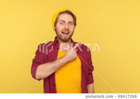 Look, advertise here. Surprised excited hipster guy in beanie hat looking at camera with happy amazed expression, pointing away at copy space, blank yellow background for commercial idea. isolated 88417269