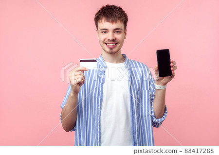 Mobile payment. Portrait of happy brown-haired man with small beard in casual shirt holding bank card and phone, smiling, satisfied with online banking. indoor studio shot isolated on pink background Mobile payment. Portrait of happy brown-haired man with small beard in casual shirt holding bank card and phone, smiling, satisfied with online banking. indoor studio shot isolated on pink background 88417288