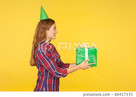 Side view of beautiful positive good natured girl with funny party cone on head giving present and saying congratulations, offering gift box, charity. indoor studio shot isolated on yellow background Side view of beautiful positive good natured girl with funny party cone on head giving present and saying congratulations, offering gift box, charity. indoor studio shot isolated on yellow background 88417335