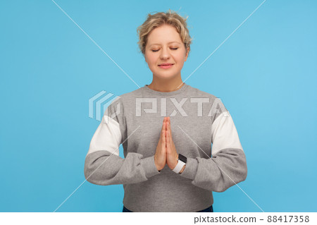 Harmony healing, yoga. Tranquil woman with short curly hair in sweatshirt doing namaste gesture and praying with closed eyes, meditation techniques. indoor studio shot isolated on blue background 88417358
