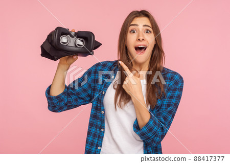 Innovative technology and cyberspace concept. Portrait of amazed gamer girl in checkered shirt pointing at virtual reality glasses, shocked by vr headset. studio shot isolated on pink background 88417377