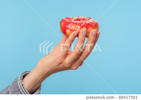 Closeup of female hand holding delicious glazed red donut with sprinkles, showing tasty doughnut, sweet confectionery high in glucose and calories. indoor studio shot isolated on blue background Closeup of female hand holding delicious glazed red donut with sprinkles, showing tasty doughnut, sweet confectionery high in glucose and calories. indoor studio shot isolated on blue background 88417381