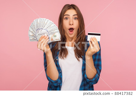 Portrait of surprised pretty girl in shirt holding credit card and dollar banknotes, surprised by lot of money, shocked by big bank loan for shopping. indoor studio shot isolated on pink background 88417382
