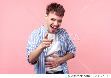 Portrait of excited happy brown-haired man with small beard and mustache in casual striped shirt pointing at camera, laughing loud at really good joke. indoor studio shot isolated on pink background 88417403