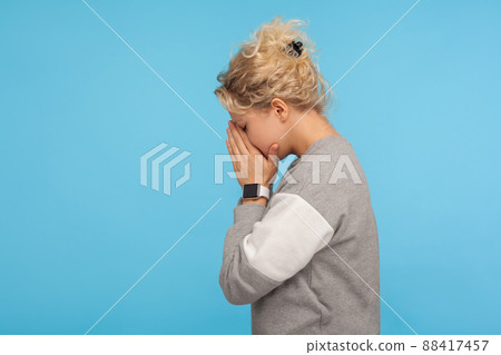 Side view of depressed woman with short curly hair in casual sweatshirt hiding face down in hands and crying, hard going through loss or defeat, feeling sorrow. studio shot isolated on blue background 88417457