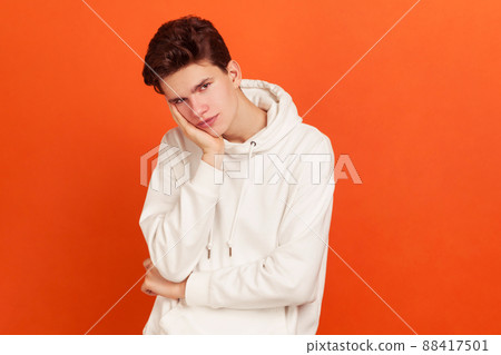 Exhausted lonely teenager in white casual hoodie looking at camera with disinterest, bored young man lost interest in life. Indoor studio shot isolated on orange background Exhausted lonely teenager in white casual hoodie looking at camera with disinterest, bored young man lost interest in life. Indoor studio shot isolated on orange background 88417501