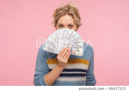 Portrait of positive woman with curly hair in sweater holding dollar banknotes, peeking out of money while hiding face, looking surprised by big profit. indoor studio shot isolated on pink background Portrait of positive woman with curly hair in sweater holding dollar banknotes, peeking out of money while hiding face, looking surprised by big profit. indoor studio shot isolated on pink background 88417525