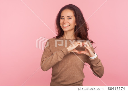 Portrait of attractive young woman with brunette hair making heart shape with hands and smiling friendly to camera, showing symbol of love and care. indoor studio shot isolated on pink background 88417574