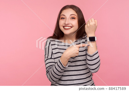 Hurry up. Portrait of positive woman in striped sweatshirt pointing smartwatch on her wrist and looking at camera with toothy smile, worried about late time. studio shot isolated on pink background Hurry up. Portrait of positive woman in striped sweatshirt pointing smartwatch on her wrist and looking at camera with toothy smile, worried about late time. studio shot isolated on pink background 88417589