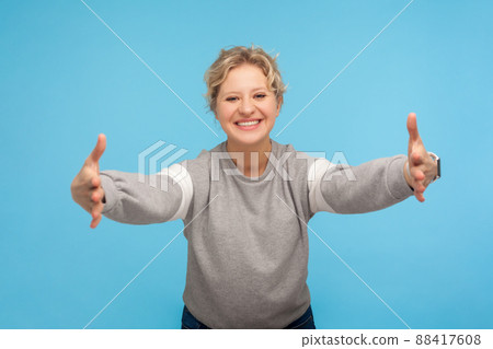 Free hugs, come here. Happy kind hearted woman with curly hair in sweatshirt opening her hands wide to give warm welcome, friendly hospitable smile. indoor studio shot isolated on blue background 88417608