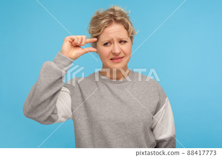 Need some more. Displeased disappointed woman with short curly hair in sweatshirt showing minimum sign with fingers, small size, little bit gesture. indoor studio shot isolated on blue background 88417773