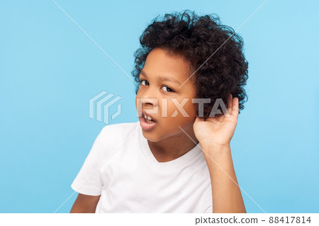 What. I can't hear you. Portrait of attentive curious little boy with curly hair holding hand near ear and listening carefully intently to what you say. indoor studio shot isolated on blue background What. I can't hear you. Portrait of attentive curious little boy with curly hair holding hand near ear and listening carefully intently to what you say. indoor studio shot isolated on blue background 88417814