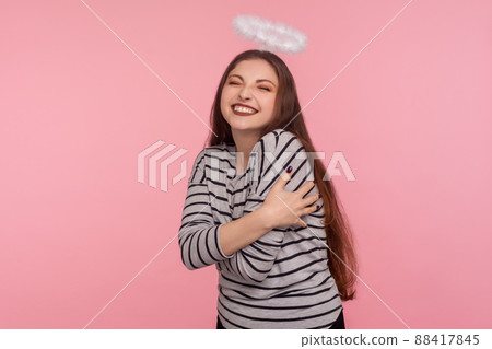 I love myself. Portrait of optimistic attractive happy woman with holy nimbus over head embracing herself and smiling carefree, positive self-esteem concept. studio shot isolated on pink background I love myself. Portrait of optimistic attractive happy woman with holy nimbus over head embracing herself and smiling carefree, positive self-esteem concept. studio shot isolated on pink background 88417845