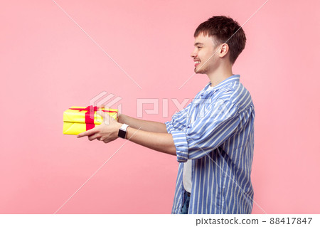 Take it. Portrait of positive generous brown-haired man with small beard in casual shirt giving present to the left side, congratulating on birthday. indoor studio shot isolated on pink background 88417847