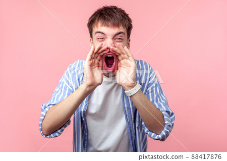 Announcement. Portrait of aggressive furious brown-haired man in casual striped shirt keeping hands near wide open mouth and screaming, shout bad news. indoor studio shot isolated on pink background Announcement. Portrait of aggressive furious brown-haired man in casual striped shirt keeping hands near wide open mouth and screaming, shout bad news. indoor studio shot isolated on pink background 88417876