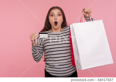 Wow credit for purchase. Portrait of amazed shocked woman holding big shopping bag and bank card, showing packages with blank space for advertise text. indoor studio shot isolated on yellow background 88417887