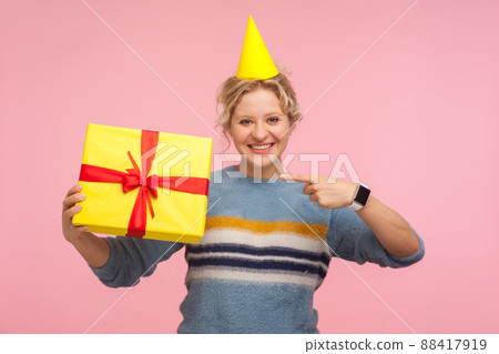 Look at my present. Portrait of happy woman with funny cone hat on head and in warm sweater pointing at holiday gift and smiling joyfully, showing birthday surprise. indoor studio shot pink background 88417919