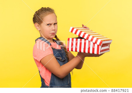 Portrait of upset child opening gift box, little girl in denim overalls looking at camera with disappointed face, almost crying, upset about bad present. studio shot isolated on yellow background 88417921