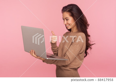Portrait of pleased young woman in casual sweater holding laptop and showing thumbs up while talking on video call, having online conference using computer. studio shot isolated on pink background 88417958