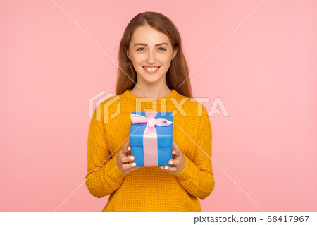 Portrait of beautiful cheerful red hair girl in sweater standing holding wrapped gift box and looking at camera joyfully with smile, festive mood. indoor studio shot isolated on pink background Portrait of beautiful cheerful red hair girl in sweater standing holding wrapped gift box and looking at camera joyfully with smile, festive mood. indoor studio shot isolated on pink background 88417967