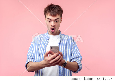 Portrait of amazed brown-haired man with small beard and mustache in casual striped shirt reading message in cellphone, astonished about shocking news. indoor studio shot isolated on pink background Portrait of amazed brown-haired man with small beard and mustache in casual striped shirt reading message in cellphone, astonished about shocking news. indoor studio shot isolated on pink background 88417997