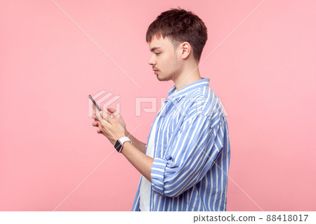 Side view of serious brown-haired man with small beard in casual shirt holding smartphone, browsing mobile internet or making call, typing message. indoor studio shot isolated on pink background 88418017