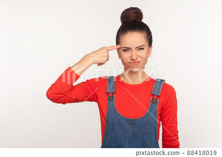 Crazy cuckoo mind. Portrait of girl with hair bun in denim overalls showing stupid gesture and looking with displeased annoyed face, accusing insane idea. studio shot isolated on white background Crazy cuckoo mind. Portrait of girl with hair bun in denim overalls showing stupid gesture and looking with displeased annoyed face, accusing insane idea. studio shot isolated on white background 88418019