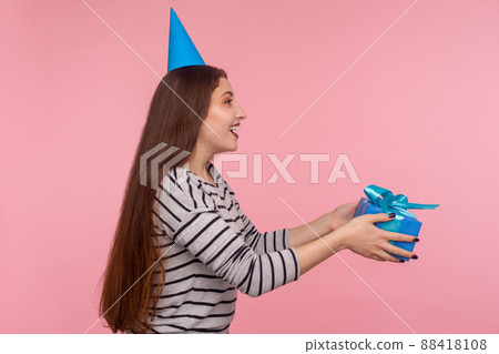 Side view, girl with party cone hat giving birthday gift and smiling, sharing present, donating and offering surprise in box, congratulation on anniversary. indoor studio shot isolated pink background 88418108