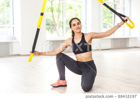 Portrait of happy beautiful young sports woman is doing TRX training, sitting on the floor and holding fitness straps while taking break. Wotkout in gym, looking at camera, indoor, window background 88418197