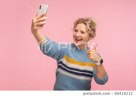 Portrait of excited pretty woman with curly hair in sweater showing thumbs up gesture and taking selfie, doing like sign with finger up while making video call. studio shot isolated on pink background 88418212