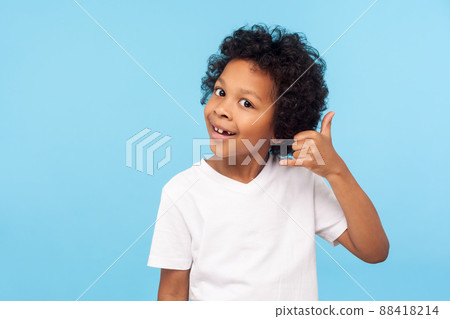 Call me. Portrait of funny little boy holding fingers shaped like telephone near head, communicating by phone, looking at camera with playful smile. indoor studio shot isolated on blue background 88418214